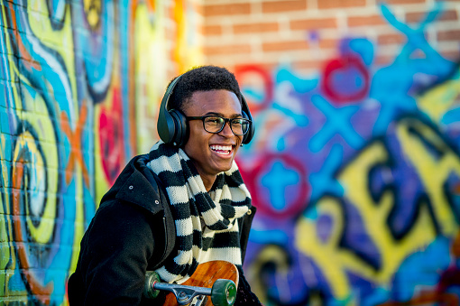 A boy of African descent is sitting in front of a wall with graffiti on it. He is wearing a trendy jacket and scarf. He is smiling while holding a skateboard and listening to music with headphones.