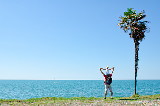 Father with son on shoulders stands back against the backdrop of tall palm, sea and blue sky