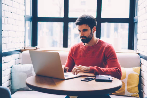 istock Skilled male programmer working on freelance concentrated on remote job while sitting in coffee shop, bearded man reading information about business startups browsing on laptop during free time in cafe 904837696