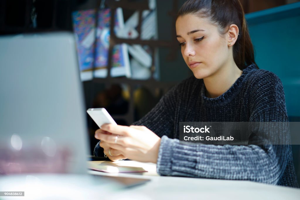 Concentrated female hipster sharing multimedia files in social networks using wireless connection and smartphone, serious young student checking email box texting messages on telephone in coworking Advice Stock Photo