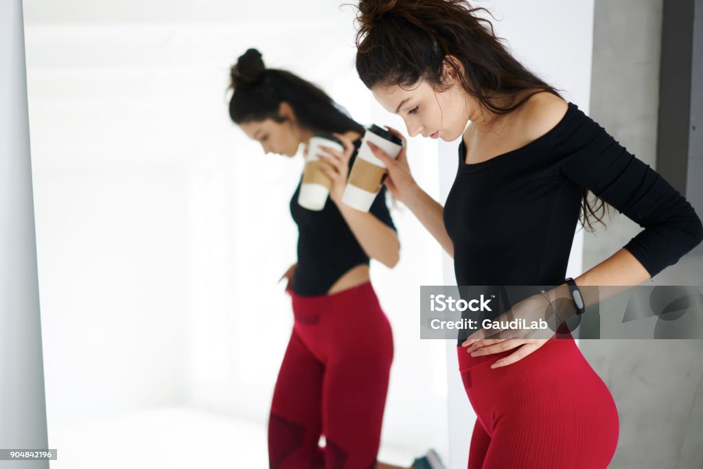 Attractive brunette female checking results of active healthy lifestyle after hard workout dressed in trendy training session, slim girl with perfect body shape resting on break looking at reflection Athlete Stock Photo