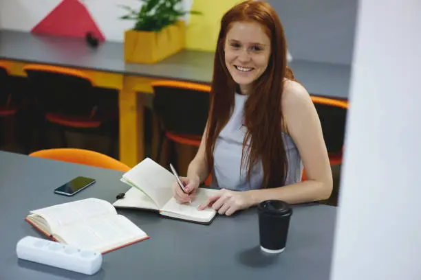 Photo of Portrait of cheerful young woman with gorgeous red hair sitting at table and rewriting some information from book for preparing to exams,smiling journalist noting plan of studying in notebook indoors
