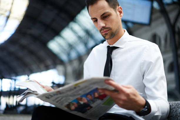 Concentrated businessman in white formal shirt analyzing information from morning press while waiting for public transport getting to job, confident entrepreneur reading newspaper on train station