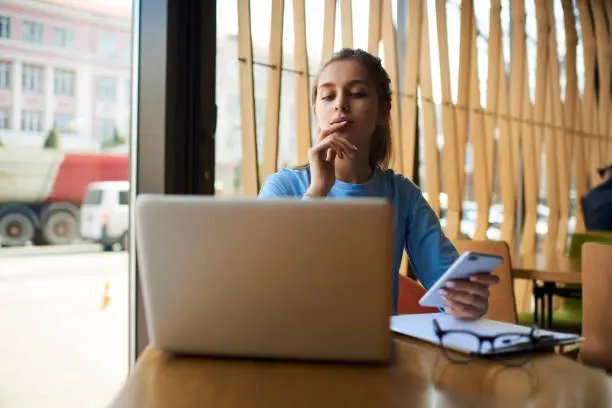 Photo of Concentrated talented student dressed in casual outfit carefully reading information on websites via macbook computer.Young hipster girl holding smartphone while looking at  modern laptop  in cafe