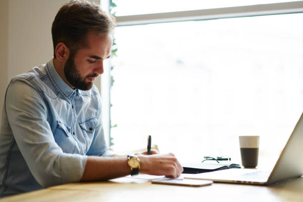 istock Handsome young businessman writing notes while planning his work day sitting at tble in office interior. Entrepreneur doing paperwork 904869260