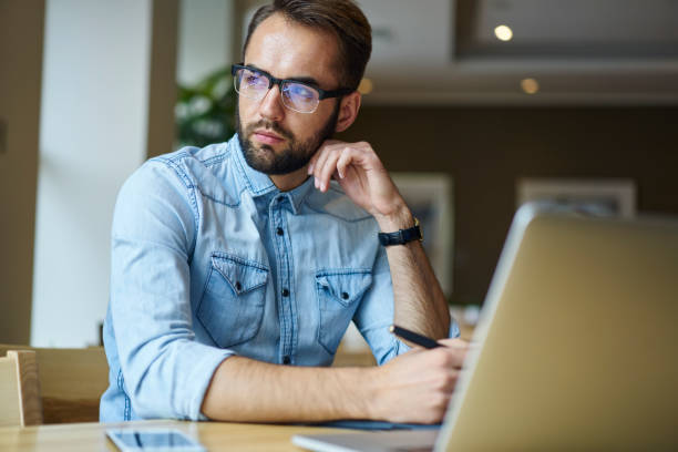istock Handsome thoughtful bearded journalist in trendy eyeglasses looking away and thinking sitting at laptop computer in coffee shop.Pensive confident male entrepreneur in spectacles working remote 904869398