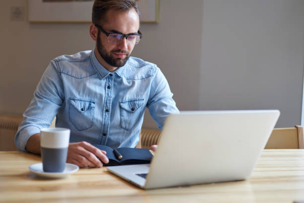 istock Young bearded manager in optical spectacles working remote at laptop computer sitting in coffee shop interior.Concentrated businessman in eyeglasses watching video on modern computer indoors 904871418