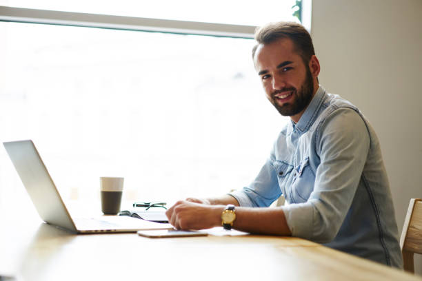 istock Portrait of cheerful bearded graphic designer working remote at modern laptop computer sitting indoors with tasty coffee while looking at camera and smiling.Advertising area for your text message 904871470