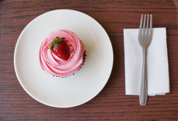 A photo of colorful cream strawberry cupcake decorated with strawberry topping and fork on white plate on wooden background, close up