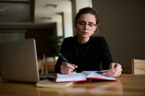 istock Concentrated professional copywriter thinking about creative ideas for new article and recording main details into notepad.Serious cute author in eyeglasses planning working process in library 904880934