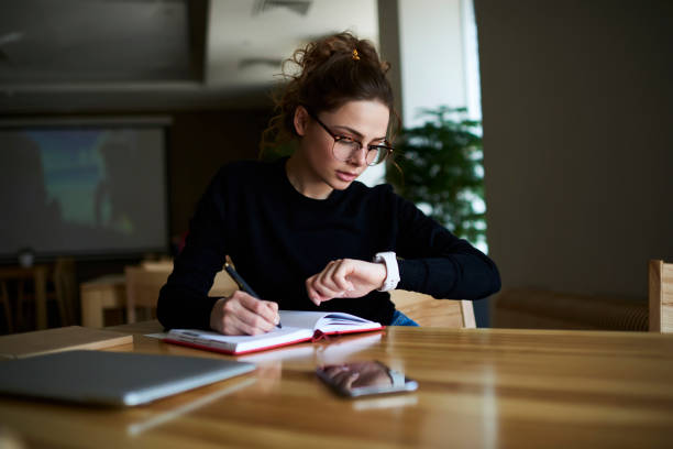 Talented female writer in stylish eyeglasses noting some information in notepad while looking on alarm watch to managing time for organization of working process sitting in university indoors talentoso escritor femenino en elegantes anteojos observando cierta información en el bloc de notas mientras se mira en el reloj de alarma para la gestión de tiempo para la organización del proceso de trabajo en la universidad en el interior