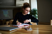 Talented female writer in stylish eyeglasses noting some information in notepad while looking on alarm watch to managing time for organization of working process sitting in university indoors