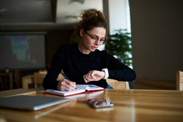 Talented female writer in stylish eyeglasses noting some information in notepad while looking on alarm watch to managing time for organization of working process sitting in university indoors Talented female writer in stylish eyeglasses noting some information in notepad while looking on alarm watch to managing time for organization of working process sitting in university indoors time management stock pictures, royalty-free photos & images