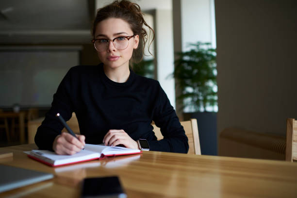 istock Portrait of smart student in eyeglasses writing checklist in notepad while looking at camera.Talented young woman with optical spectacles noting some records in notebook sitting in university interior 904880984