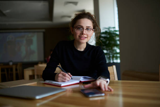 istock Portrait of intelligent student with optical spectacles writing some records in textbook while sitting at table with laptop computer in coworking space and looking at camera during exam preparation 904881172