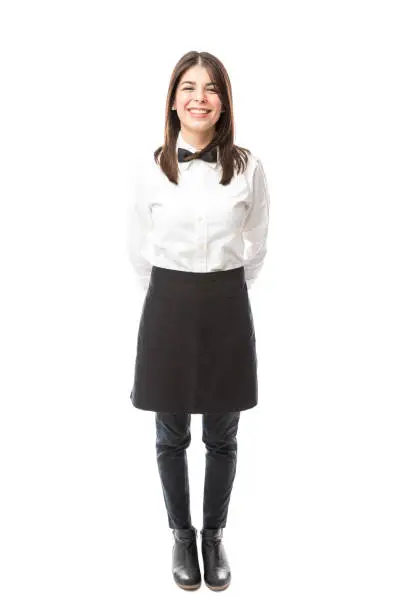 Full length view of a beautiful Hispanic waitress wearing an apron and a bowtie and smiling;