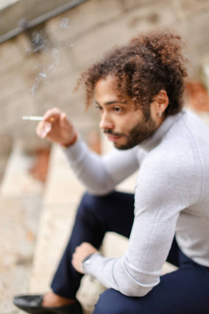 Handsome  male person smoking and coughing, sitting on steps.  Young boy wears grey turtleneck sweater, watch and dark pants. Concept of propaganda healthy lifestyle and quit smoking.
