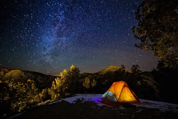 Camping in a Tent Under the Stars and Milky Way Galaxy - Scenic landscape with hils and mountains with orange glowing tent in dark with view of night sky with Milky Way and bright stars.