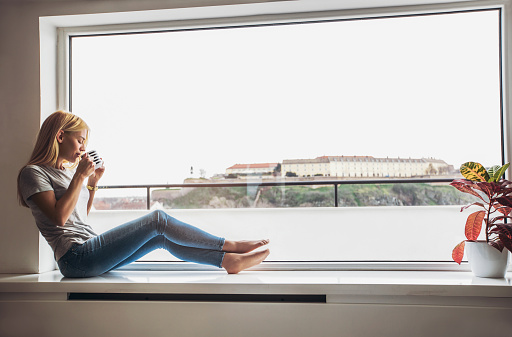 Beautiful young woman drinking coffee and looking through window while sitting at windowsill at home