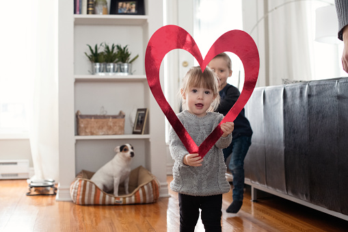 Celebrating love at Valentine's day with two lovely children of 18-23 months and 4-5 years old, sister and brother, holding together a heart shape. They are standing up in the kitchen and look through the red heart shape. A Jack Russell dog is in the back ground. Photos was taken in Quebec Canada.A