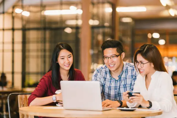 Photo of Young Asian college students group or coworkers using laptop computer together at cafe or university. Casual business, freelance work, coffee break meeting, e-learning or e-commerce activity concept