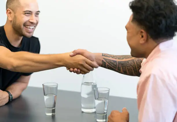 Two Polynesian business men in a city office setting in New Zealand shake hands smiling