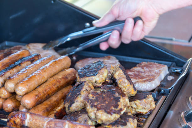 Outdoor barbecue scene shows a hand using tongs to flip several pieces of meat on a sizzling grill. Char marks, smoke, and glowing embers create a warm, savory dining moment.