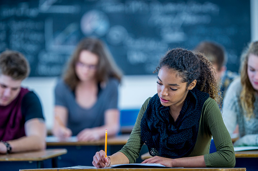 A multi-ethnic group of high school students are indoors in their classroom. A girl of African descent is in focus, and she is writing a test with her pencil.