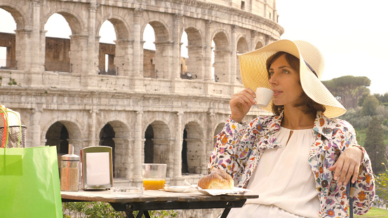Happy young woman tourist drinking coffee and juice with cornetto at the table outside a bar restaurant in front of the Colosseum in Rome. Elegant beautiful dress with large hat and colorful shopping bags