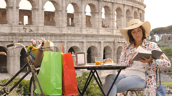 Young woman tourist reads a book sitting at the table outside a bar restaurant in front of the Colosseum in Rome with coffee, juice and cornetto. Elegant beautiful dress with large hat and colorful shopping bags