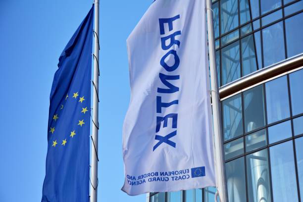 European Union flag and Frontex banner flying outside a glass office building against blue sky.