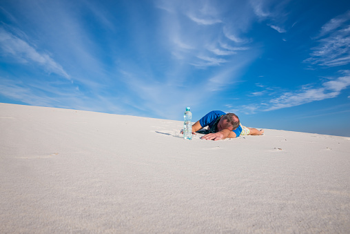 Tired man, lost in the desert, lies exhausted in the sand next to a bottle of water, suffering from thirst, on the background of blue sky. Wide angle.