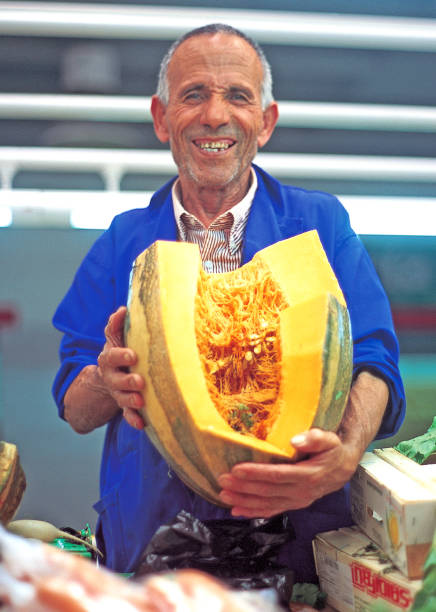 In the fruits a vegetables market of Melilla City a vendor smiles while holding with his two hands a very large Pumpkin .