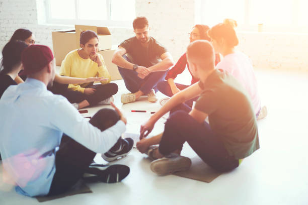 Young talented architects talking about interior planning and organization of  working process in new office sitting indoors with flare sun.Male and female employees discussing details of cooperation