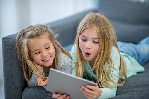 Two Caucasian sisters are indoors in their house. They are sitting on the couch and watching a movie on a tablet. One girl is very happy, and the other is shocked.