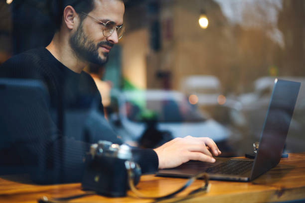 Bearded entrepreneur male in glasses dressed in black sweater reading news checking email and chatting with friends in social network using wireless internet connection at wooden table in coffee shop Bearded entrepreneur male in glasses dressed in black sweater reading news checking email and chatting with friends in social network using wireless internet connection at wooden table in coffee shop cd writer photos stock pictures, royalty-free photos & images