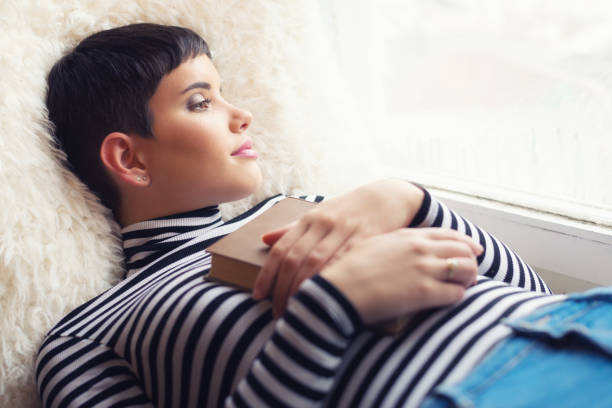 istock Woman reading book beside window 905169678