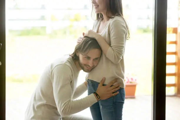 Photo of Happy man putting ear to pregnant wife belly at home