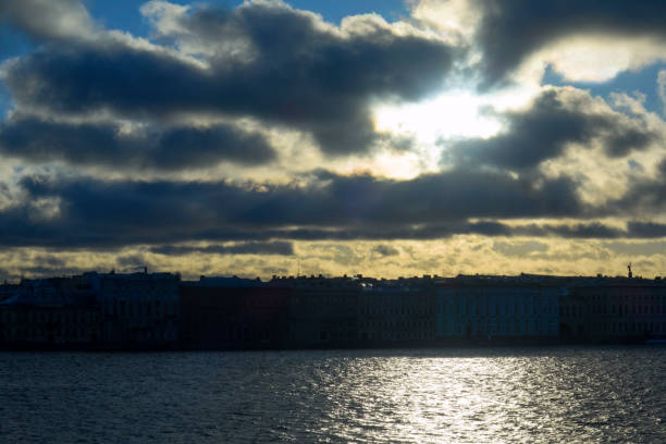 The sun and clouds in the sky, the reflection of the sun in a wide river. A big city in the shade.