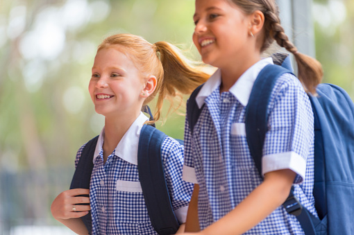Schoolgirls in uniform with back pack. She is with her friend at school. They are very happy smiling and holding hands