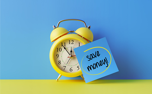 Yellow alarm clock with a blue post it note attached over bright blue background. Save money writes on post it note. Reminder concept. Horizontal composition with copy space.