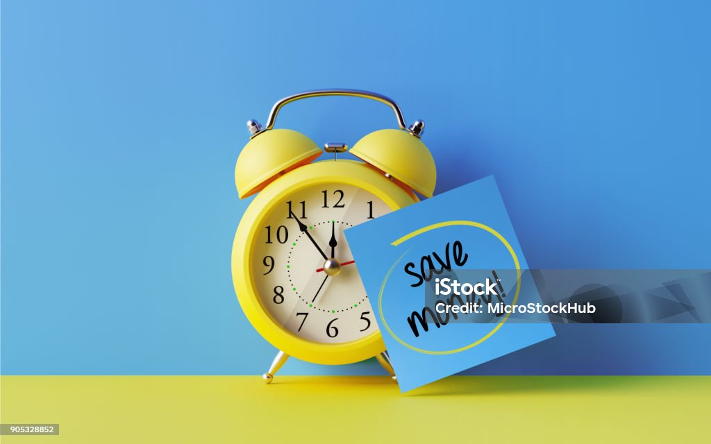 Alarm Clock And A Blue Post It Not Over Blue Background Yellow alarm clock with a blue post it note attached over bright blue background. Save money writes on post it note. Reminder concept. Horizontal composition with copy space. Savings Stock Photo