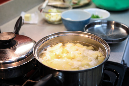 Food preparations for potato salad or mashed potatoes for holiday dinner.  Potatoes are boiling in the pot on the stove.  Other ingredients are in bowls awaiting inclusion in the meal.
