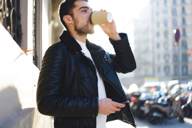 istock Good looking hipster guy dressed in trendy clothing enjoying energetic beverage while walking at urban setting.Handsome bearded male drinking aroma coffee during strolling outdoors in city on sunny day 905358470