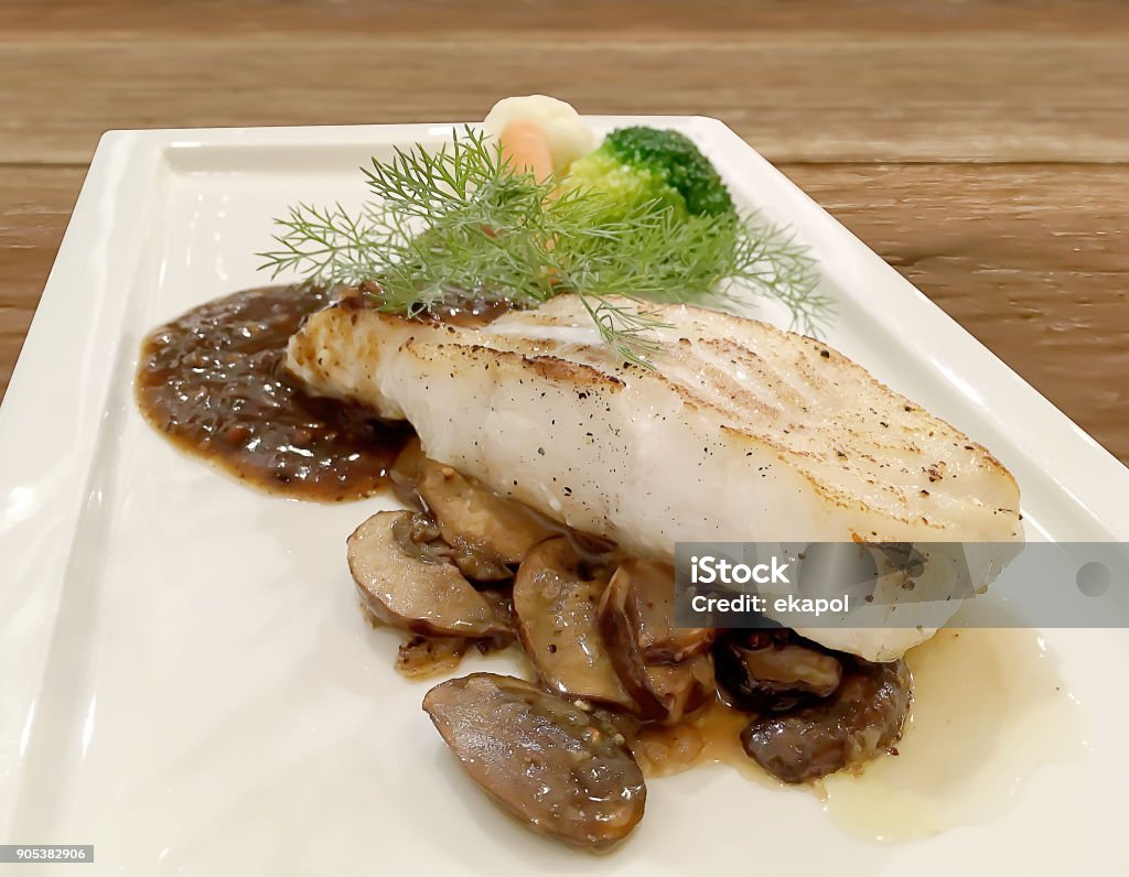Selective focus closeup on Grilled Snapper fish stake with mushroom Sauce and fresh green vegetable in white plate on wooden table. Close-up Stock Photo