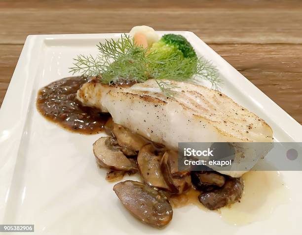 Selective Focus Closeup On Grilled Snapper Fish Stake With Mushroom Sauce And Fresh Green Vegetable In White Plate On Wooden Table Stock Photo - Download Image Now