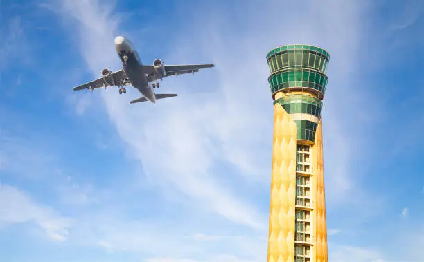 Airplane flying over air traffic control tower and blue sky.