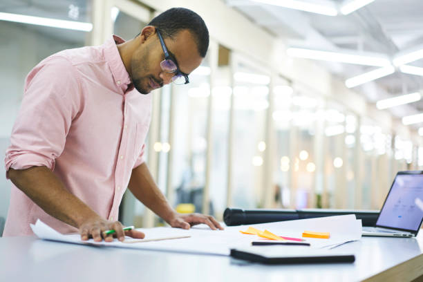 istock Male web designer looking at printed sketch of skyscraper for putting data information into application on laptop computer with mock up screen connected to wifi while creating model of interior 905431734