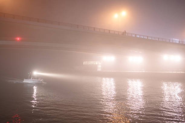Night city in fog, people walk on the bridge. Boat floats on the channel lighting the way a bright spotlight. Dubai, United Arab Emirates.