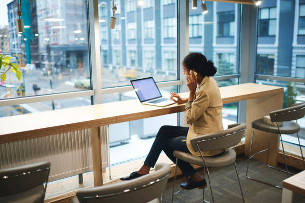 jungen afroamerikanischen exekutive macht remote job wird in geschäftsreise sitzt am tisch im coworking-raum im gespräch am telefon mit unternehmer mit laptop-computer mit mock-up bildschirm mit wifi - gutsangestellter stock-fotos und bilder